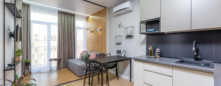 Open space moderno e luminoso con cucina compatta, tavolo da pranzo, piccolo divano e ampie finestre con tende chiare. Arredi minimal che ottimizzano lo spazio in un appartamento di dimensioni ridotte.